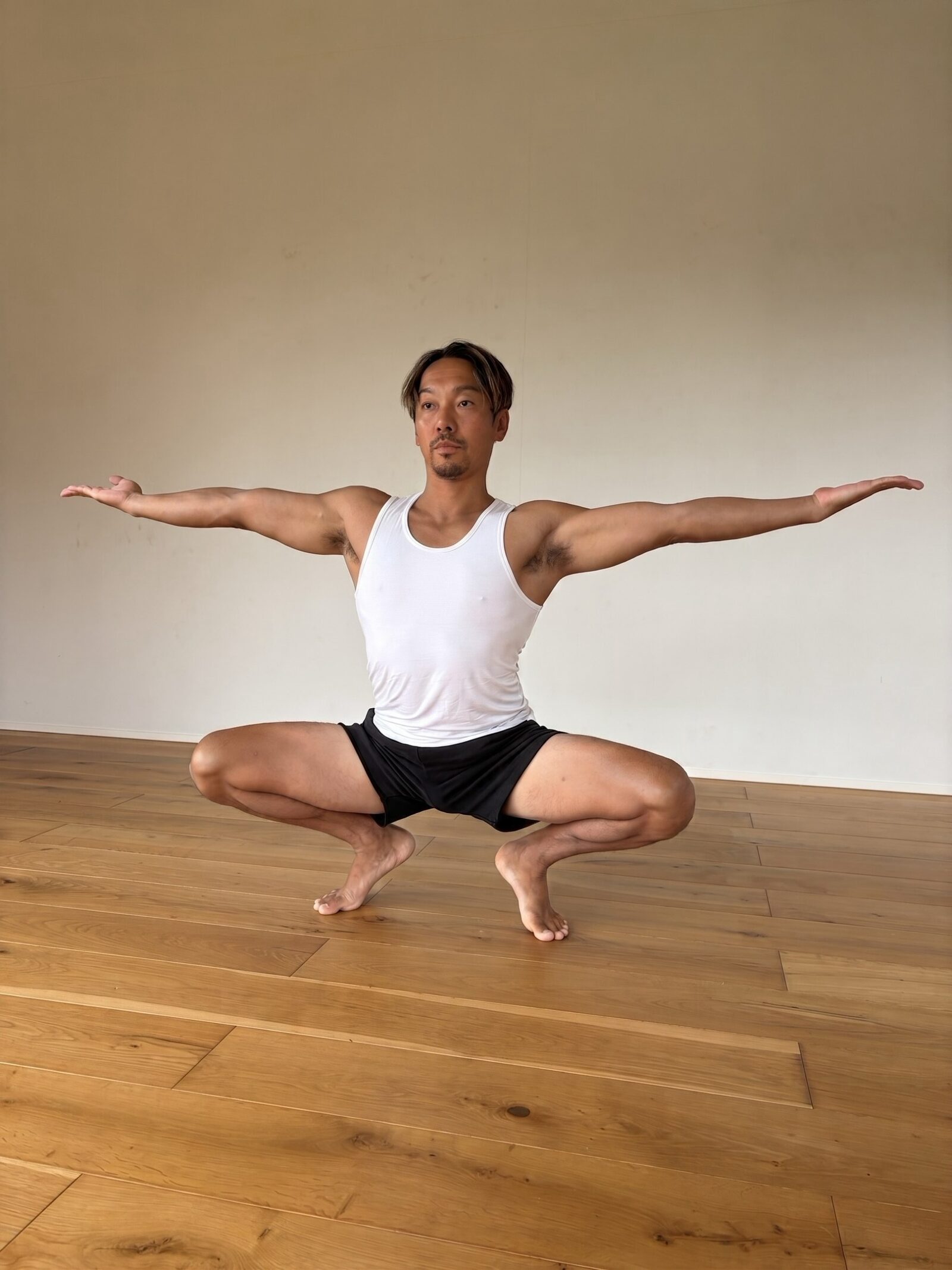 A deep squat with both arms stretched out to the sides on a wooden studio floor